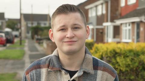 Frankie Williams with short brown hair smiling into the camera. He's wearing a blue and cream checked shirt. He's stood outside in a residential area and in the background are houses, a hedgerow and parked cars.