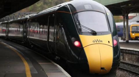 Yellow and dark green train travelling towards the camera with two red lights lit on its front and a wiper on the windscreen