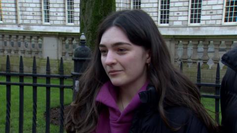 A young woman with long brown hair is wearing a purple hoody and black coat. She is standing in front of a set of black railings and a white bricked building.