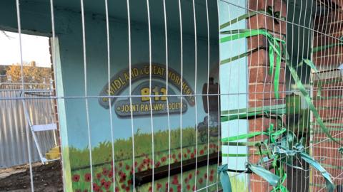 Bus shelter wrapped in construction barrier wire with green ribbons tied to the wall