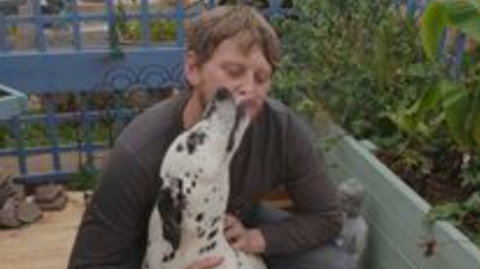 Ryan Blackwell is in a garden hugging a Dalmatian dog that is licking his face. He has a dark long-sleeved T-shirt on