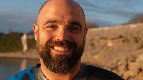 Jason with dark hair and beard, wearing a blue and black swimming wetsuit
