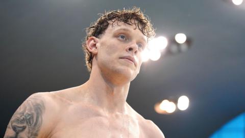Matt Richards pictured looking into the distance. His hair is wet and curly having come out of the water and he is topless, showing a lion tattoo