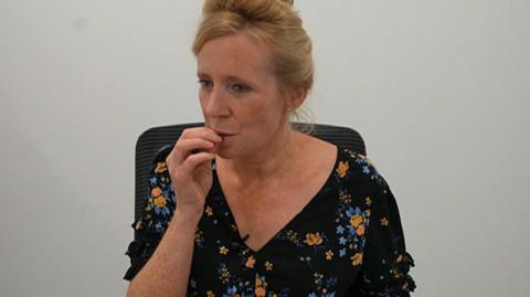 A woman with auburn hair in a top bun hair style tastes a piece of chocolate. She is sitting on a chair and is wearing a black dress, which has blue and orange flowers on it. 