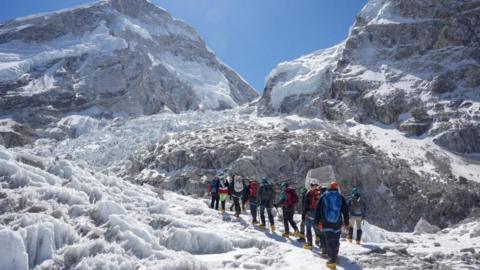 Members of an expedition team are pictured in a line at Khumbu Icefall in front of an ice and mountain backdrop with a blue sky