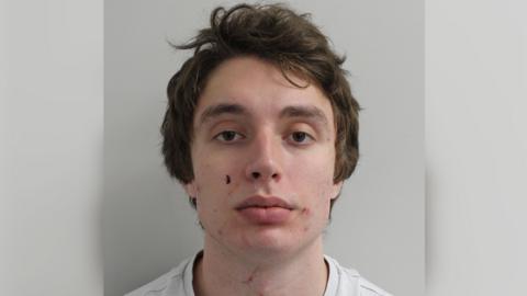 Custody image of Matvei Rumiantsev. He is a young man with short brown hair, wearing a white t-shirt. He stands with a neutral expression in front of a white wall.