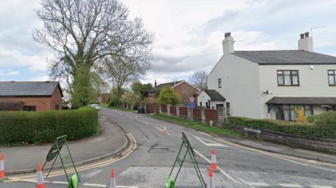 Paradise Lane pictured from Dunkirk Lane in Leyland. Paradise Lane sits between a red brick building on one corner and a light rendered two-storey house on the other. There is a large tree near the junction and 20mph signs along with a dead end symbol. A couple of houses can be seen down the street. There are roadworks at the junction with orange and white traffic cones continuing along the centre road markings of Paradise Lane. The picture was taken in 2023.