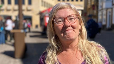 A woman in a purple dress stands in a market square on a sunny day. There is a brown bin out of focus behind her, and buildings and people out of focus.