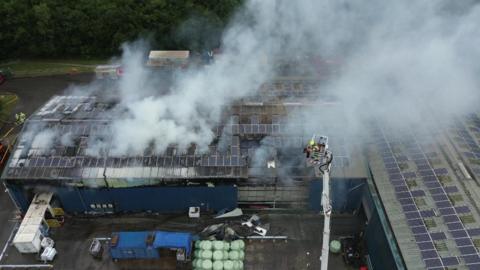 Smoke rises from a large industrial building with an aerial ladder visible with at least one firefighter on it and a number of bales of material in green wrapping on the ground.