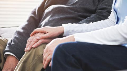 A stock image of a nurse putting her hand on an elderly gentleman's hand. They are sitting down.