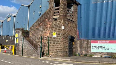 An industrial building, with blue corrugated sheeting and brick-built sections. There is a sign with the words Shrub Hill Industrial Estate in capital lettering.