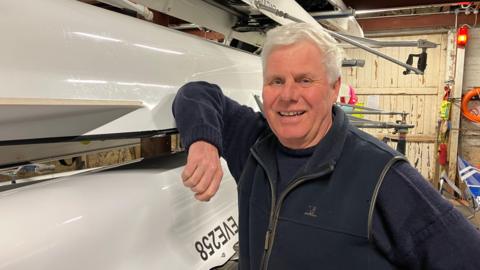 A man with white hair leans on a white rowing boat with one arm, in a storage area for boats. He is wearing a navy fleece and jumper and smiling.
