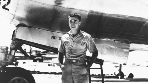A black and white photo of Hiroshima mission pilot Paul Tibbets stood in front of the shiny silver Enola Gay plane, which he flew to drop the atomic bomb on Hiroshima. In the picture he is young, has short dark hair and is wearing a raggedy shirt with the top buttons undone, holding his hands behind his back.