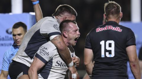 Nick Tompkins of Saracens roars with delight after scoring a try as Theo Dan jumps on his back with Newcastle no.19 Freddie Clarke having his back turned