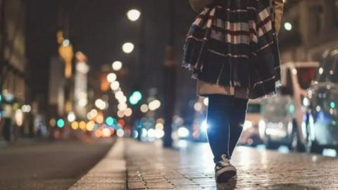 An anonymous woman walks down a street at night, with black and white sneakers, with black tights, and a beige coat.  To her left is a row of vehicles and to the right a row of buildings - both are blurred out.