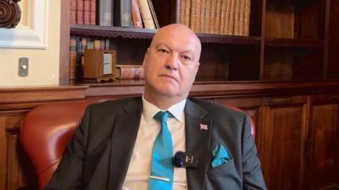 Ian Cooper sits in a maroon leather chair, with wooden panelling and a recessed bookcase behind him. He is wearing a white shirt, dark grey suit jacket, with a union flag pin badge and a clip microphone on the lapel, and a teal blue tie. He is looking into the camera with a serious expression.