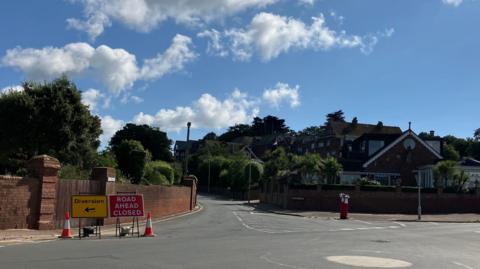 A red road ahead closed sign and yellow diversion sign with an arrow pointing left have been set up by a mini-roundabout at the junction with Maer Road in Exmouth following a sewage spill in August. It is a sunny day with a few clouds in the sky. A red letter box is on the opposite side of the pavement to the road signs.