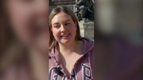Blonde woman wearing a pink jacket looks to the side with an awkward smile.