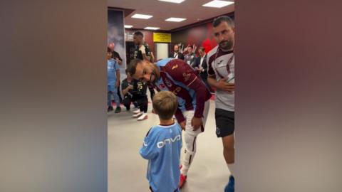 Image shows Mo Salah leaning down to talk to little Isaac Kearney. They are in the dressing room at the club, and are both wearing the club's kit.