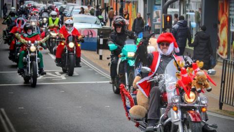 The bikers lining up the streets, dressed in Santa clothes. Their bikes are adorned with Christmas toys and tinsel. People are taking pictures of them from the sidewalk. Buster Spicer leads the ride, he is also wearing black shades. He is waving.