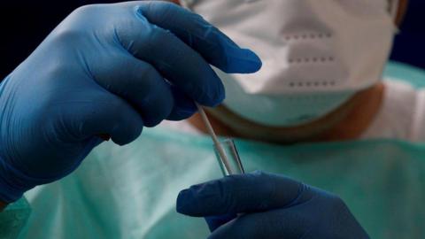 A stock image of a person wearing PPE which includes blue gloves, a white facemask and a mint-coloured bib. The person is holding a test tube