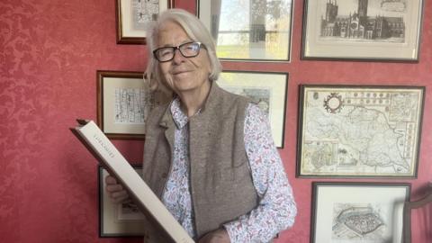 A smiling woman with short grey hair wears black rimmed glasses, a gilet, and a floral shirt. She holds a large book and stands in front of a red-wallpapered wall, which is full of old, framed maps and photographs.