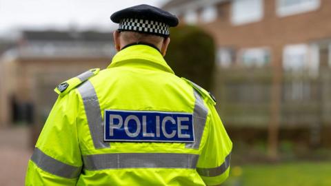 The back of a police officer, in a high-visibility neon yellow jacket with the word police on the back, is shown