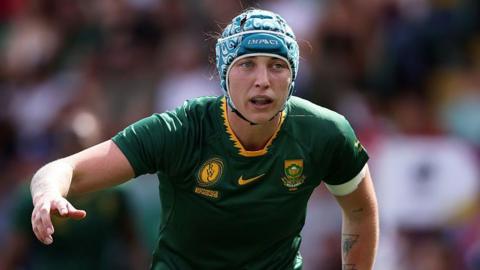 Danelle Lochner, wearing a scrum cap and South Africa kit, bent forward while standing on the pitch with her right arm stretched out to the side during the Rugby World Cup
