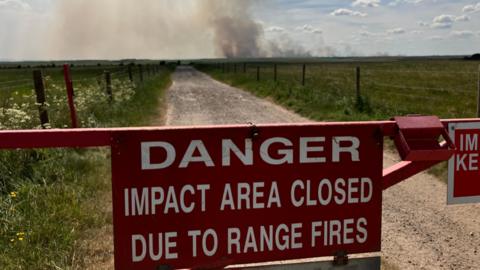 A red metal gate looking down a countryside track with large fields down either side and a large plume of smoke in the distance. Sign reads 'Danger impact area closed due to range fires'
