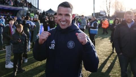 John Rooney on the pitch, smiling and posing with clenched fists, after Macclesfield's FA Cup win against Crystal Palace