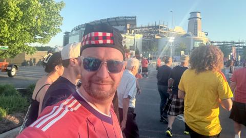 Man wears a Scotland top, hat and sunglasses, outside a sport stadium
