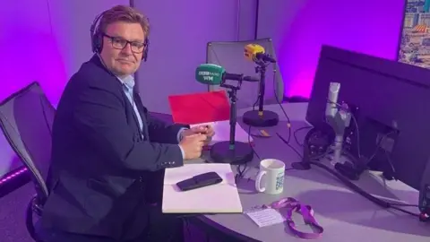 A man in a shirt and blue jacket is sitting side on to the camera, but with his head facing the camera. He is sitting by a green microphone in a radio studio that has a purple background.