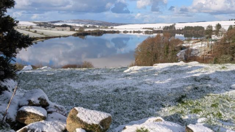 The picture shows a peaceful winter landscape with a large lake in the middle reflecting the blue sky and scattered clouds above. Surrounding the lake are gently rolling hills covered in a light layer of snow, giving the scene a crisp and bright appearance. In the foreground, there is a grassy area partially dusted with snow and a few large rocks lying near the edge. On the right side, there are clusters of leafless trees and some evergreen trees. The background stretches into distant hills, also blanketed in snow, under a sky that mixes patches of blue with dramatic white and grey clouds.