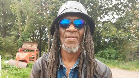 A man called Hubert Brown with dreadlocks wearing a hat and blue-tinted sunglasses looks at the camera.