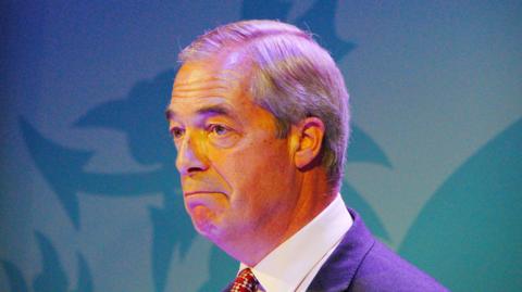 Nigel Farage stood in front of a light blue Dragon backdrop in the colours of his party. He is wearing a tie that is mostly out of shot with a suit and white shirt.