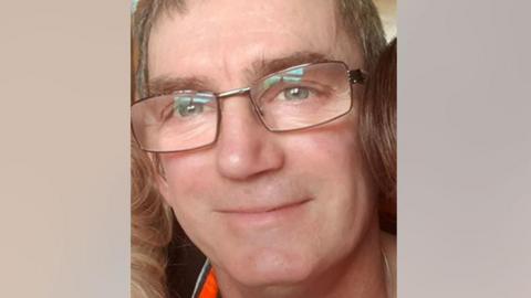A man in his 50s with short hair and glasses smiles at the camera 