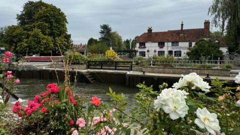 Red and white flowers can be seen in front of the River Thames, with a white building in the background under overcast skies in Sandford-on-Thames 