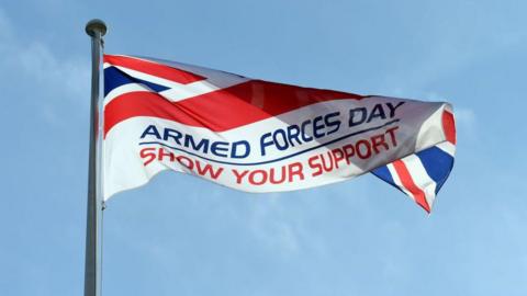 A red, white and blue flag featuring the text "ARMED FORCES DAY, SHOW YOUR SUPPORT" written in caps. It is flying from a flagpole. The sky is blue.