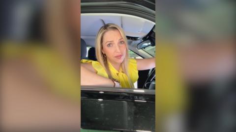 Presenter Emma Vardy looks out of the window from the driving seat of a car. She has long blonde hair and wears a yellow top