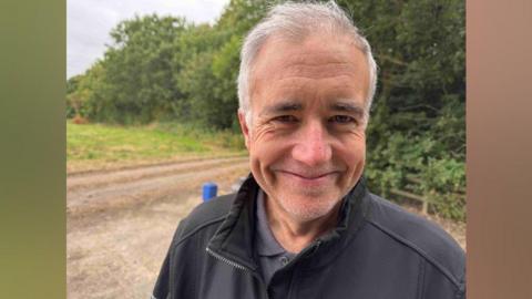 A man with grey-white hair is smiling at the camera. He has a black zip jacket on, and is standing on a gravel road with green grass and trees beyond