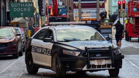 A police car with Gotham Police written on the side drives along a road. A green sign in the background says Gotham Docks in white capital letters. Film crews are in the background with lights.