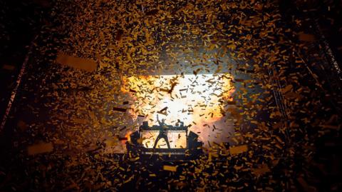 Fatboy Slim's silhouette is in the distance, his arm aloft, as he plays music at decks. Huge swirls of confetti are all around