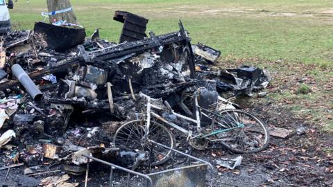 The remains of a burnt-out caravan and bike next to a patch of grass. Little of the remains are identifiable as they form a pile of black, charred rubble. 