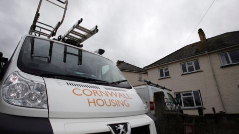 A Cornwall Housing van parked outside of some houses. The photo is taken from the front bumper of the white van looking up at it.