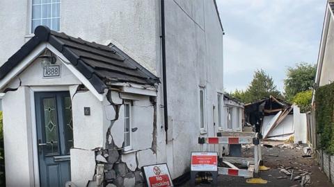 A white rendered house with a porch with a blue door that has massive cracks on it and is reduced to rubble in the right corner. To the side is a driveway with a broken garage and red and white signs warning not to smoke