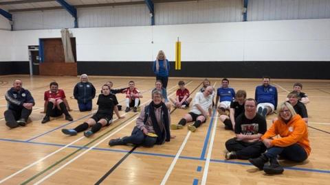 The Guernsey Dynamics Football team and coaches with some members of the swimming club sitting on the floor of a gymnasium posing for a photo