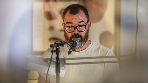 Stuart Whipps in a white t-shirt speaking into a microphone, with an indistinct projection behind him