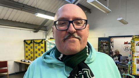 A bald man with a grey moustache and black rimmed glasses looking at the camera, he is wearing a pale green jumper and a green Plymouth Argyle football scarf. He is standing in a community centre with chairs and other furniture in the background.