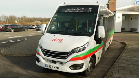 A white minibus with A2B Travel Group in red lettering on its bonnet. There are single red and green stripes from the front of the bus, running along its side. The bus destination board above the windscreen says Duxford WM Whittlesford Circular 7f, and a bus driver is visible in the driver's seat. It is parked in a layby, with a bus shelter and a car park visible in the background 