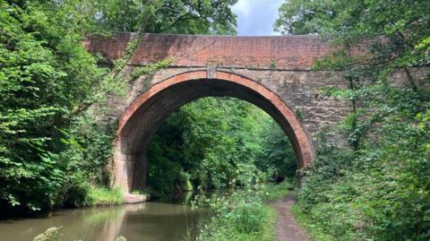 A brick bridge over the canal. A footpath and trees and bushes can be seen on the each side.